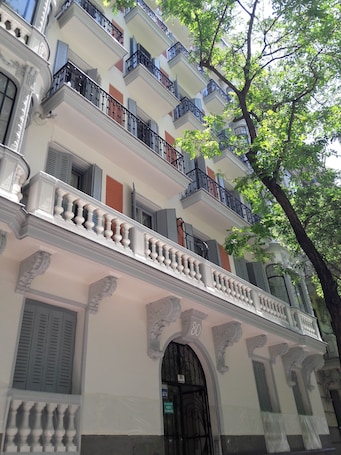 A multi-story building with balconies, a central entrance with an archway, and a tree in the foreground.