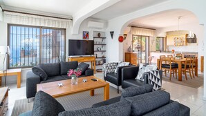 A modern living room with a sofa set, a coffee table, and a TV. There is a dining area with a table and chairs.