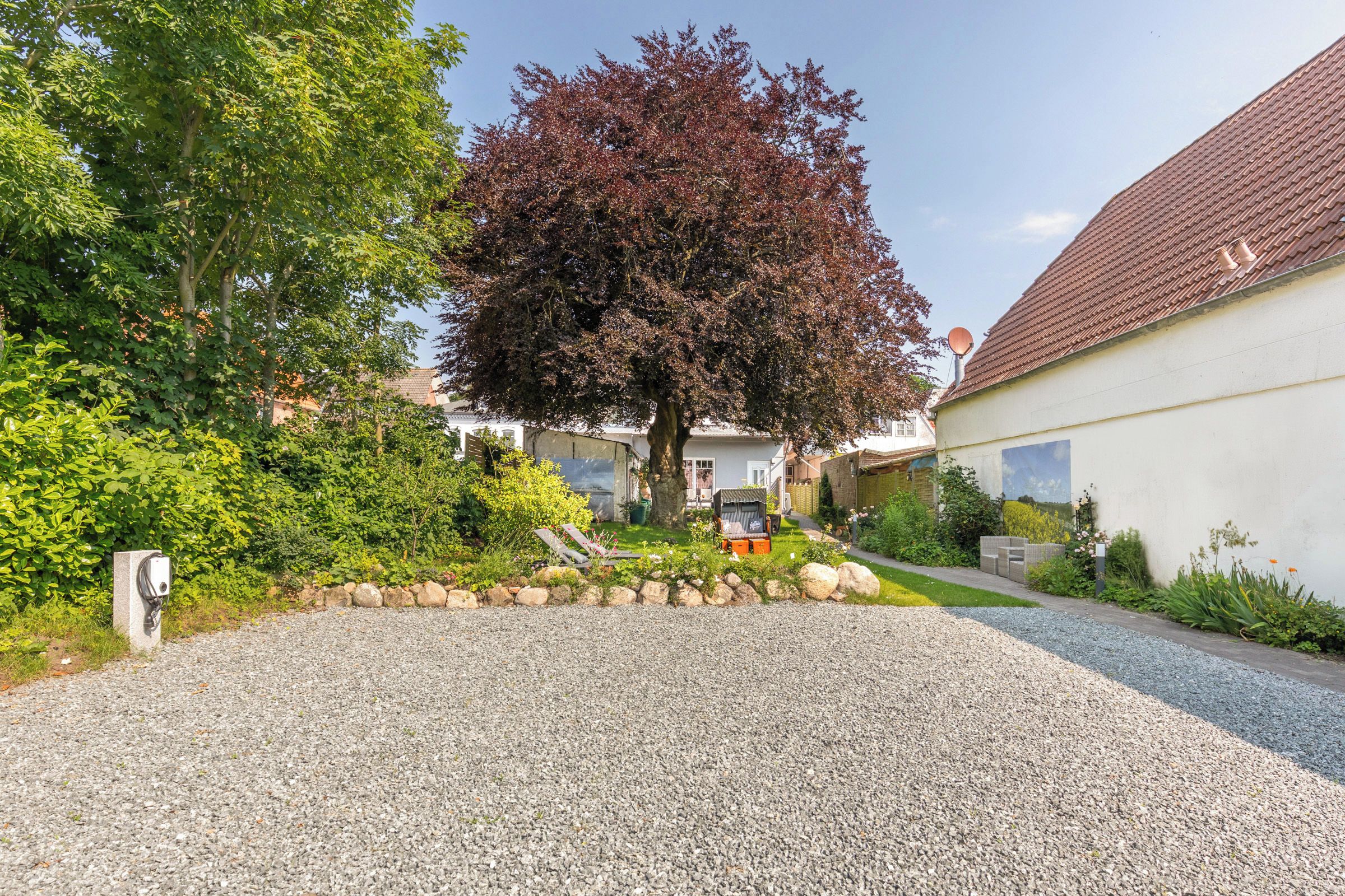 A residential driveway with a gravel surface, a tree with red leaves, and a house with a tiled roof.