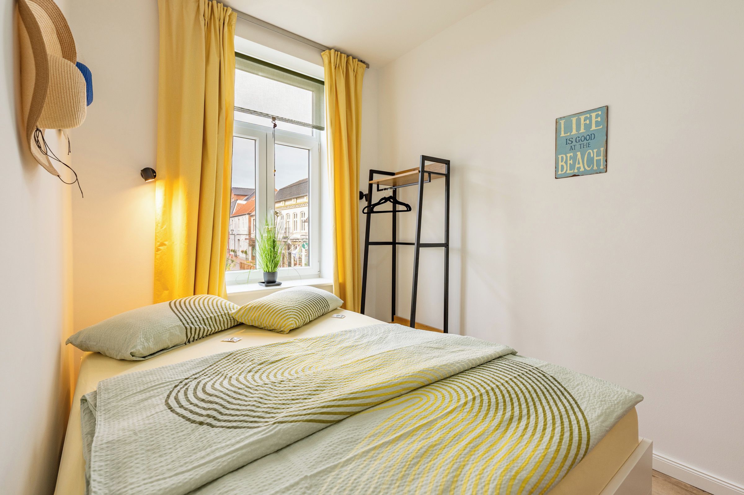 Une chambre avec un lit, des rideaux jaunes, une fenêtre donnant sur un paysage agréable, un porte-manteau fixé au mur et un panneau avec du texte.