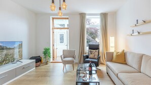 A modern living room with a sofa, armchair, and coffee table. There is a television on the left and a window with curtains. - Die Ferienwohnung "ellis Groot Stuuv" in Garding für bis zu 4 Personen (Garding)