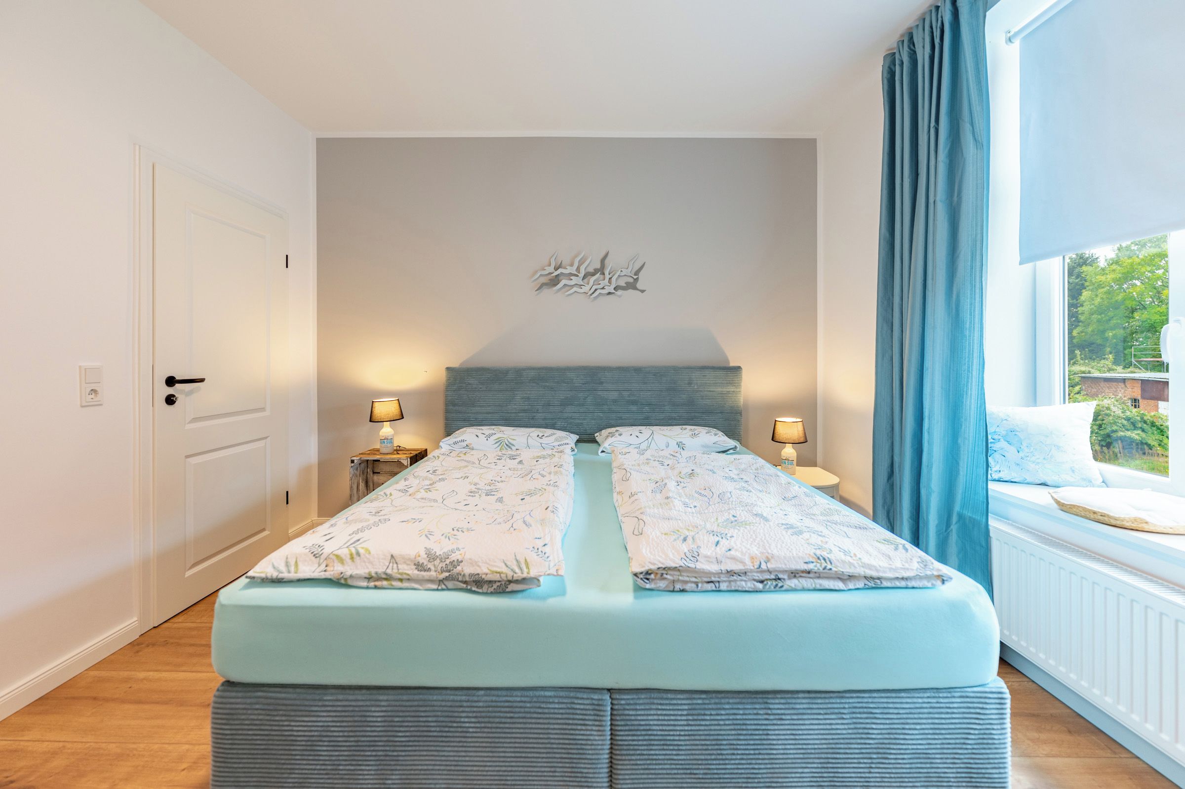 A bedroom with a bed, two bedside tables, a wall-mounted light fixture, and a window with curtains.