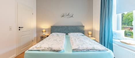 A bedroom with a bed, two bedside tables, a wall-mounted light fixture, and a window with curtains.