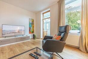 A modern living room with a flat-screen TV, a glass coffee table, a leather armchair, and a large window with curtains.