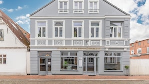 A two-story building with a grey facade and white trim. It has a central entrance with glass doors and windows on the ground floor. - Die Ferienwohnung "ellis Groot Stuuv" in Garding für bis zu 4 Personen (Garding)