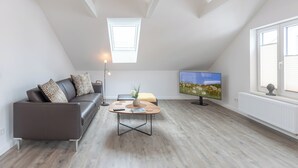 A modern living room with a sloped ceiling, a sofa, a coffee table, a TV, and a window with blinds. - Die Ferienwohnung "gretes Dackstuuv" in Garding für bis zu 4 Personen (Garding)