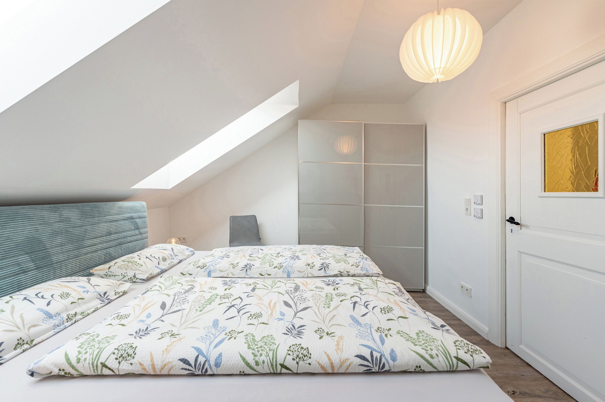 Een slaapkamer met een schuine kap, een bed met bloemenbeddengoed, een nachtkastje, een stoel en een kledingkast.