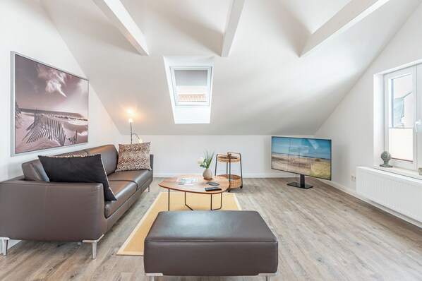 A modern living room with a brown leather sofa, a coffee table, a TV, and a window with a view of the beach. - Die Ferienwohnung "gretes Dackstuuv" in Garding für bis zu 4 Personen (Garding)