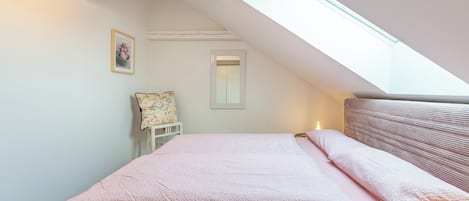 A bedroom with a sloped ceiling, a bed with pink bedding, a small bedside table, a chair, and a framed picture on the wall.