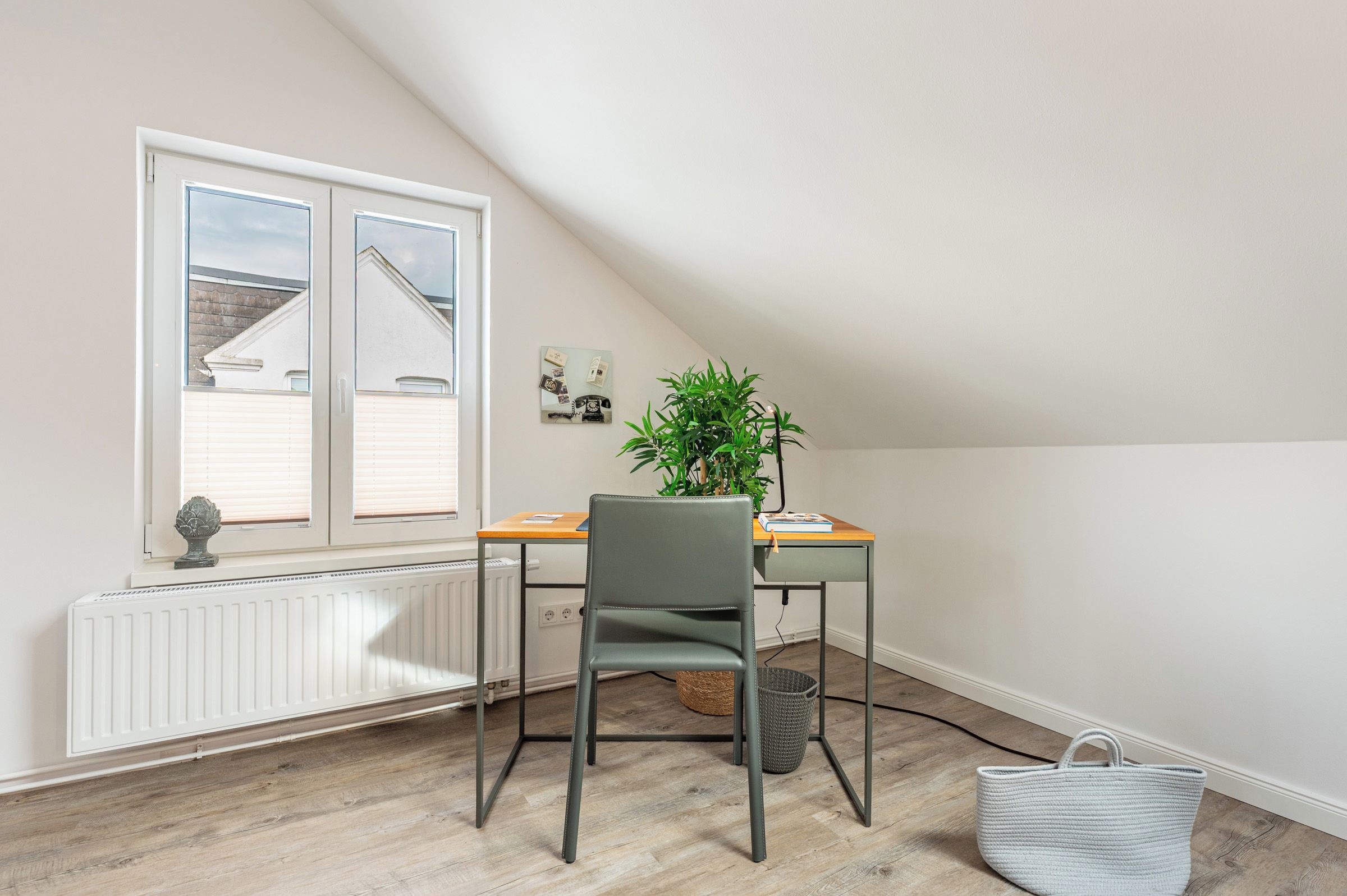 Une pièce avec un plafond en pente, un radiateur, une fenêtre avec des stores, un bureau avec une chaise, une plante en pot et un panier en osier.