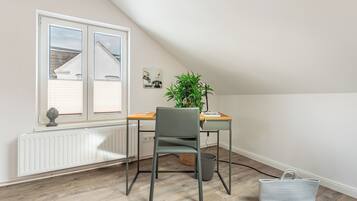 Une pièce avec un plafond en pente, un radiateur, une fenêtre avec des stores, un bureau avec une chaise, une plante en pot et un panier en osier.