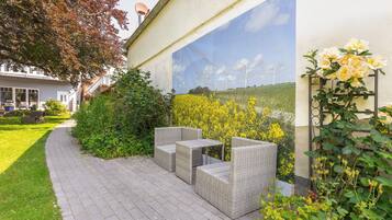 Un patio con mobiliario de mimbre, un jardín con flores amarillas y un mural con aerogeneradores en la pared.