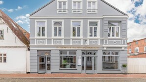Een gebouw met twee verdiepingen, een grijze gevel en witte details. Het heeft een centrale ingang met glazen deuren en ramen op de begane grond.