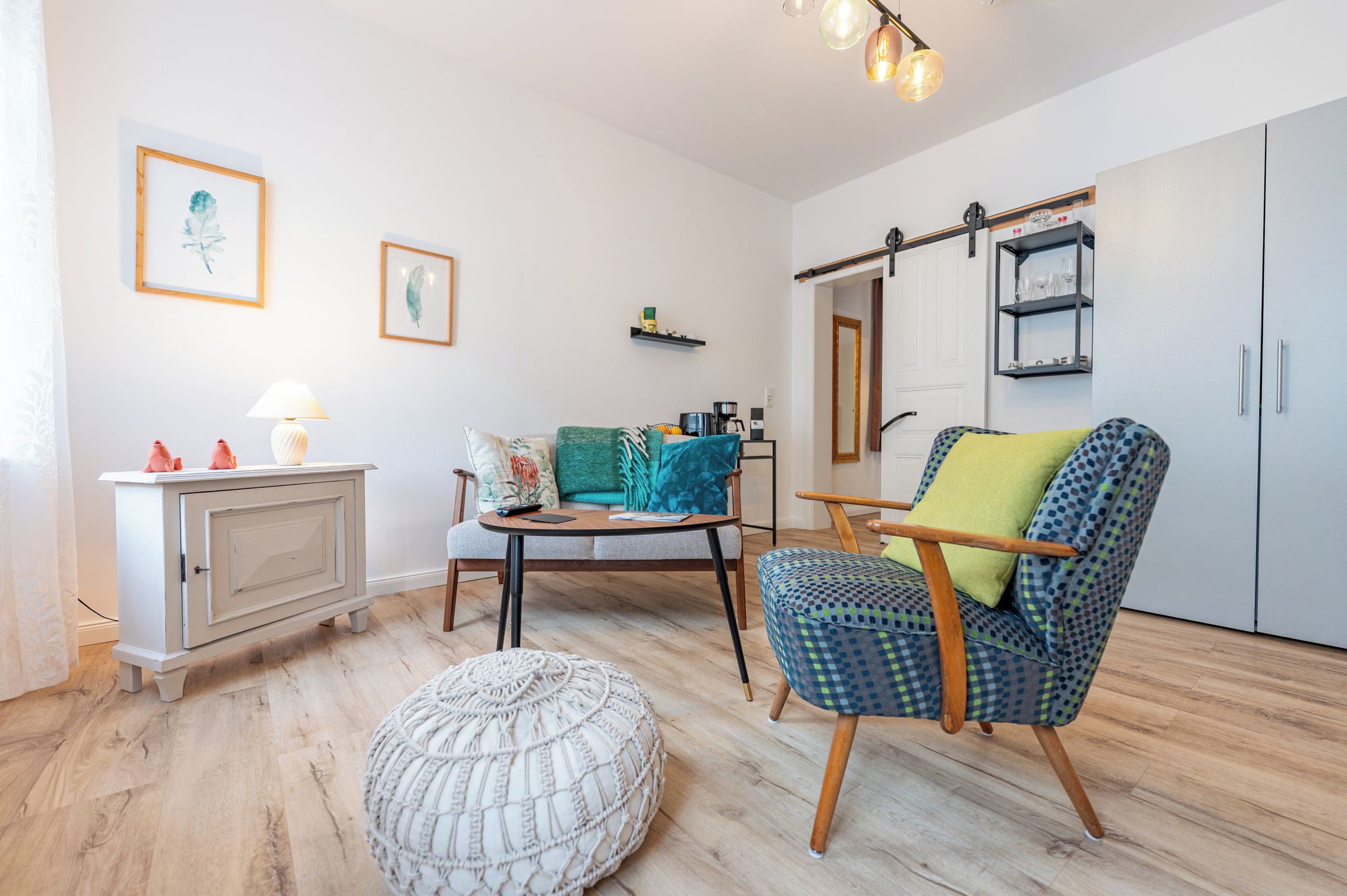 Ein gemütliches Wohnzimmer mit Sofa, Sessel, Hocker, einem kleinen Tisch mit Lampe und einem Schrank.