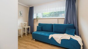 Una habitación con un sofá azul, sillas blancas, una ventana con cortinas, un radiador y un cuadro enmarcado en la pared.