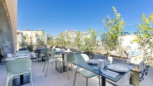 Un patio al aire libre con mesas y sillas, rodeado de plantas en macetas y edificios al fondo.