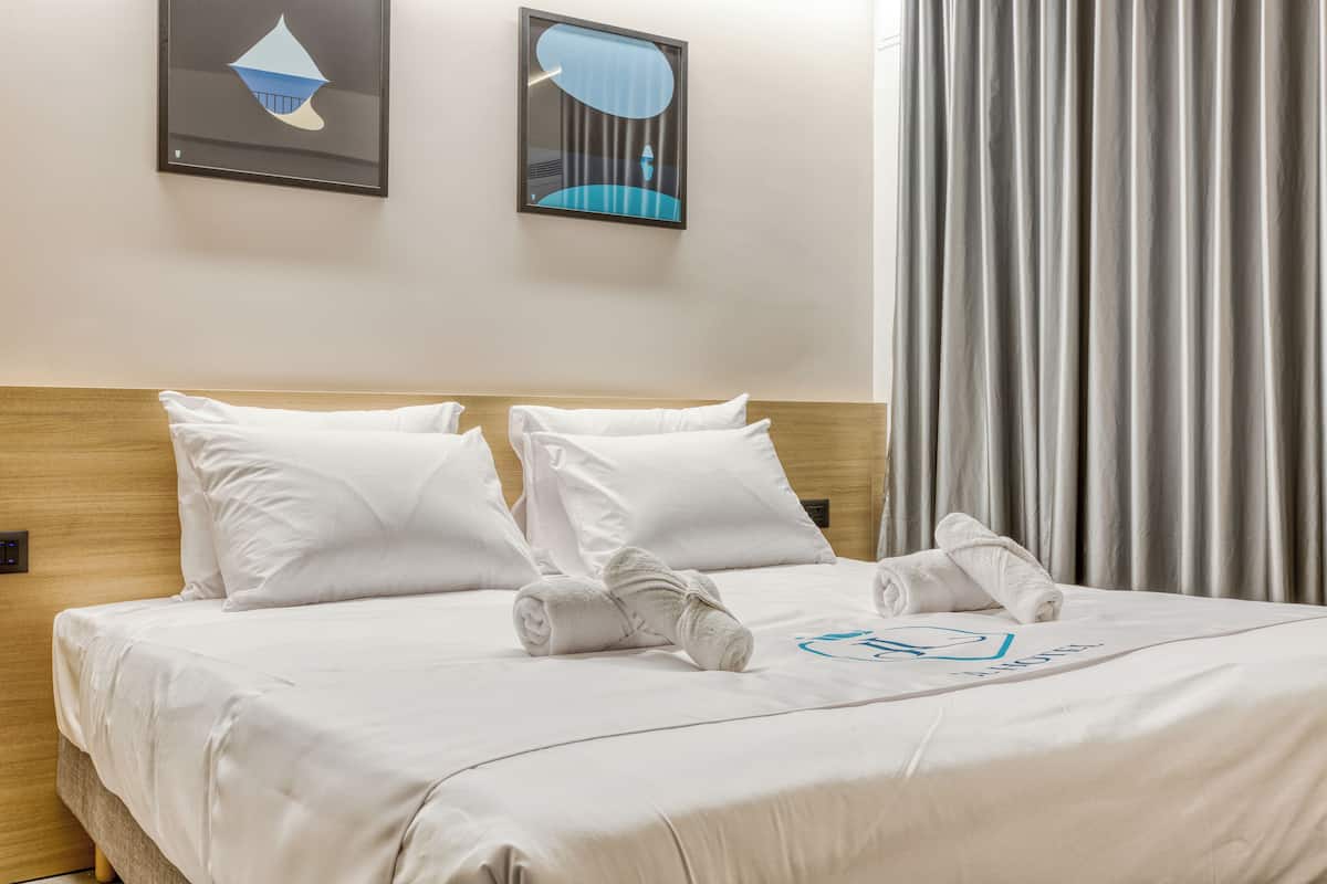 A neatly made bed with white linens and towels, a wooden headboard, and two abstract paintings on the wall.