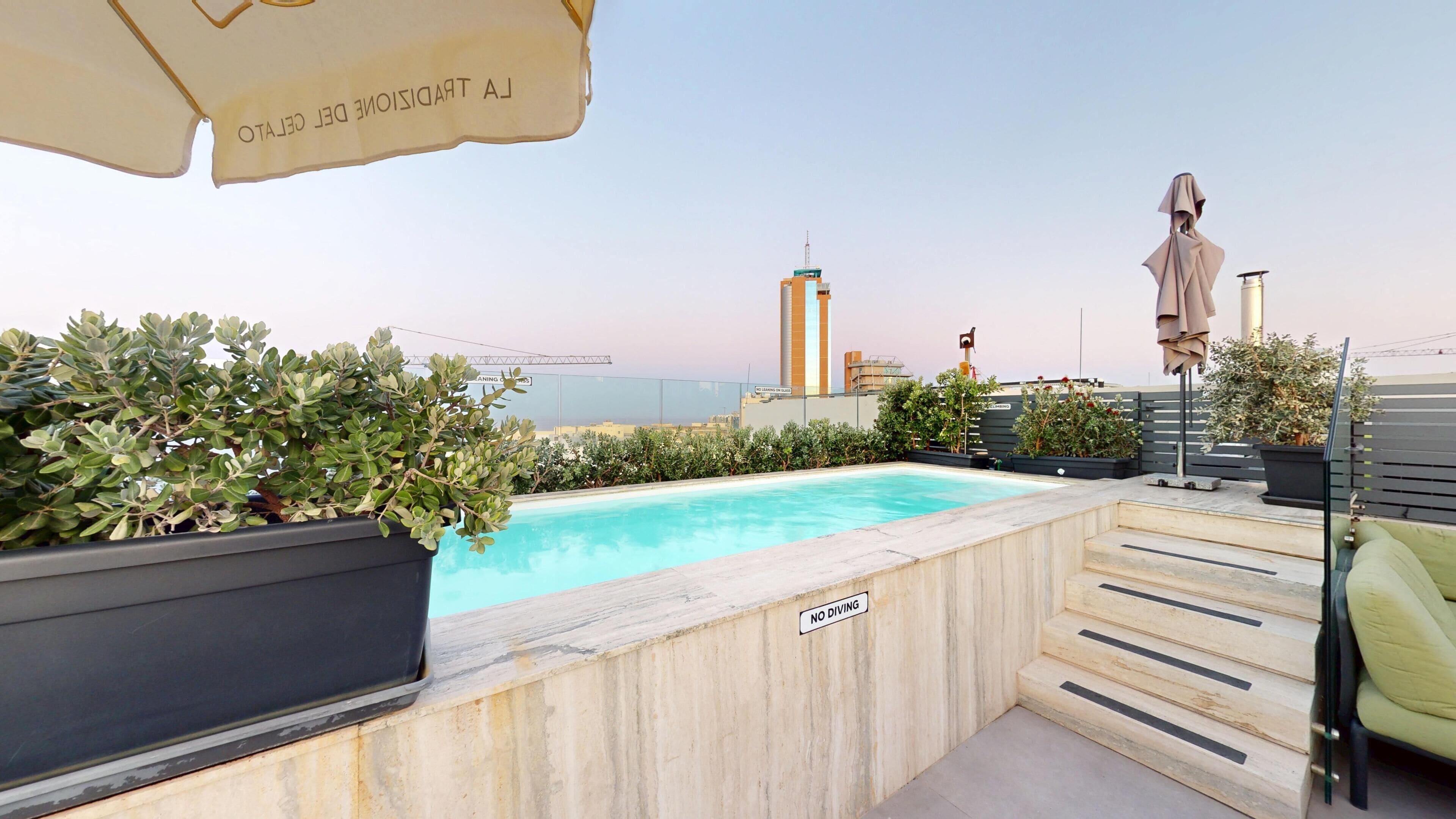 A rooftop pool with a 'NO DIVING' sign, potted plants, a staircase, and a view of the city skyline.