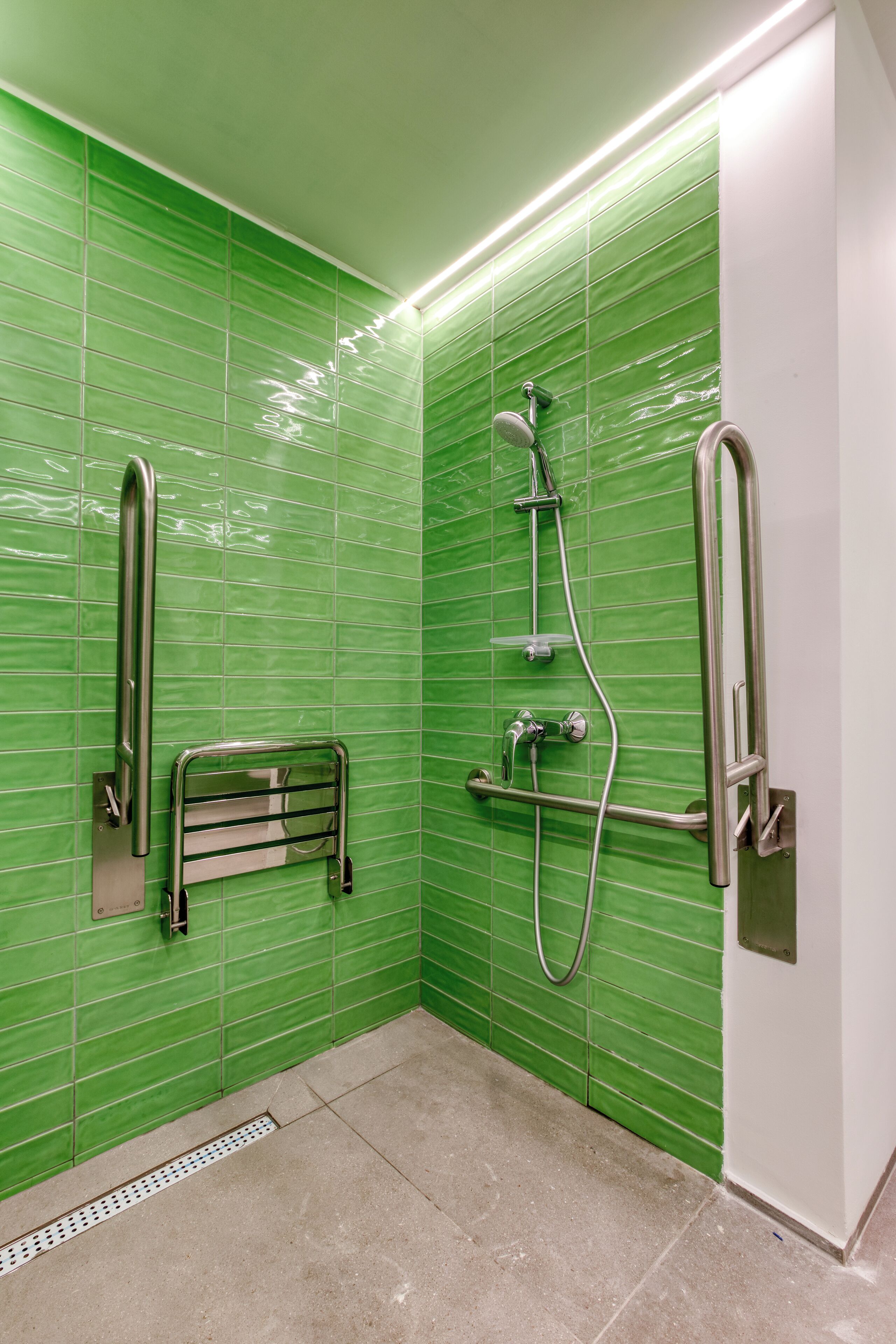 A bathroom with green tiled walls, a shower with a handheld showerhead, a grab bar, and a toilet with a safety grab bar.