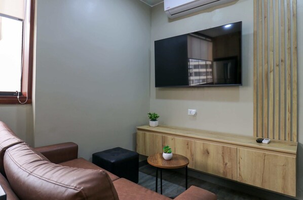 Una sala de estar moderna con un sofá de cuero marrón, una pequeña mesa redonda de madera con una planta, un reposapiés negro, un mueble de consola de madera con un televisor montado encima, y una ventana con persianas.