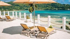 A beachside seating area with lounge chairs and umbrellas overlooking a turquoise sea and green hills. - The Bay Seychelles Boutique Hotel and Apartments (Mahé Island)