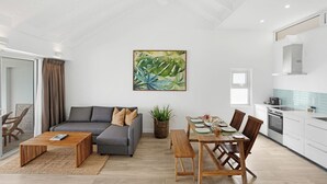 A modern living room with a grey sofa, wooden coffee table, and a dining area with a wooden table and chairs. A large painting hangs on the wall, and there is a potted plant in the corner. - The Bay Seychelles Boutique Hotel and Apartments (Mahé Island)