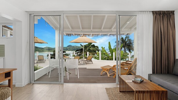 A modern living room with a sliding glass door leading to an outdoor patio area with seating and umbrellas. - The Bay Seychelles Boutique Hotel and Apartments (Mahé Island)