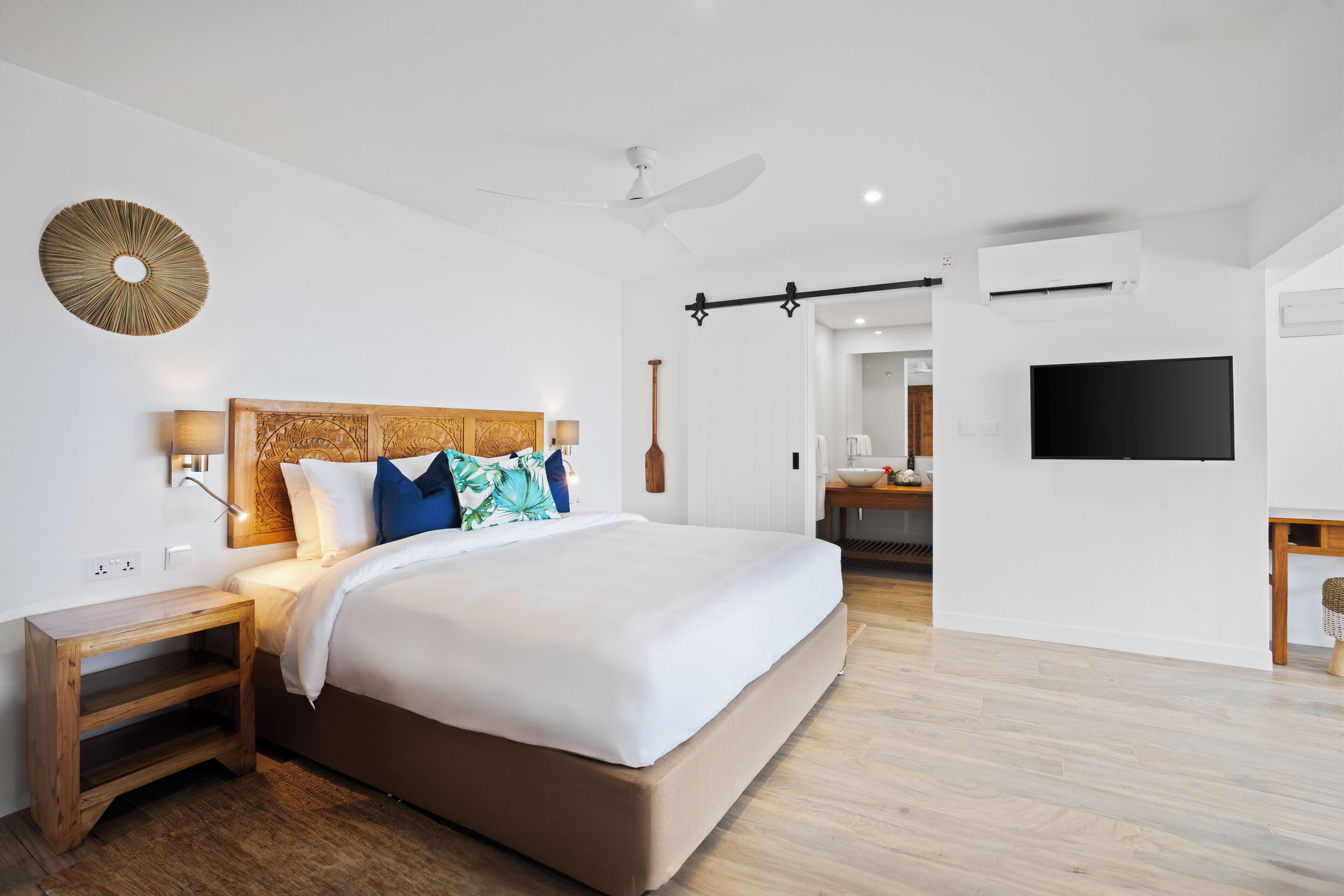 A modern hotel room with a large bed, wooden headboard, bedside table, and a flat-screen TV mounted on the wall.