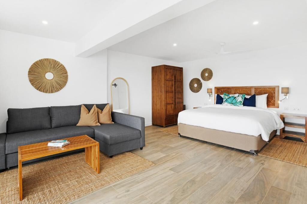 A modern hotel room with a grey sofa, a wooden coffee table, a large bed with a wooden headboard, and a wardrobe.