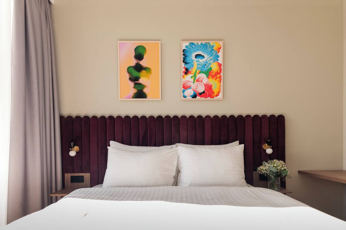 Une chambre d’hôtel avec un lit, deux lampes fixées au mur, une tête de lit sombre et deux œuvres d’art encadrées.