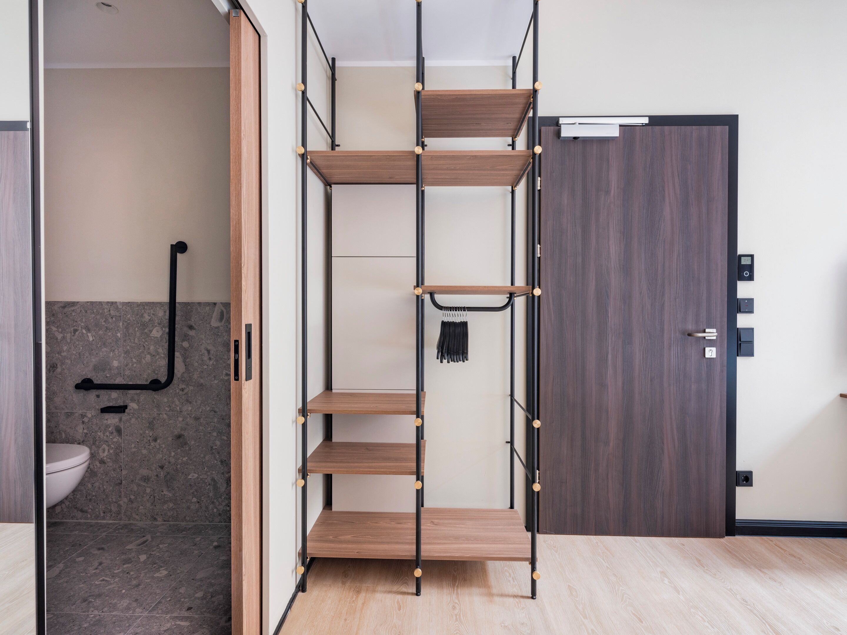 A room with a wooden floor, a built-in shelving unit with multiple shelves, a door with a digital lock, and a towel rack.