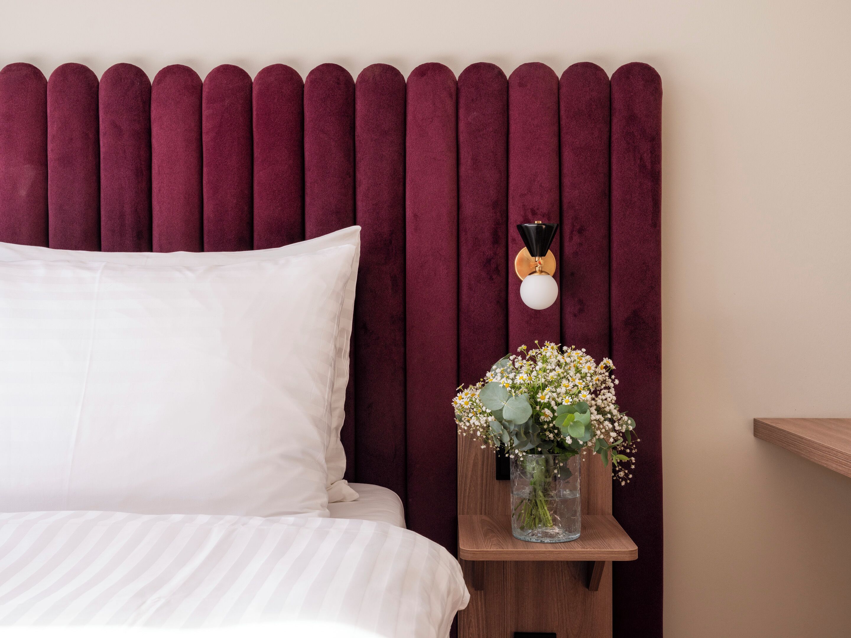 A bed with white bedding, a dark red headboard, a bedside table with a vase of flowers, and a wall-mounted light fixture.