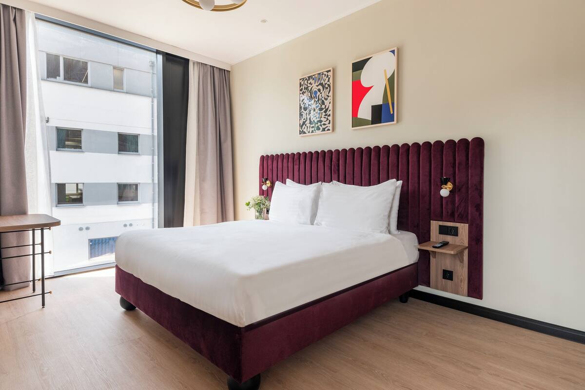 Une chambre d’hôtel avec un grand lit, une table de chevet, une commode, une lampe fixée au mur et une fenêtre avec des rideaux.