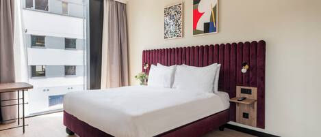 Una habitación de hotel con una cama grande, una mesita de noche, una lámpara de pared y una ventana con cortinas.