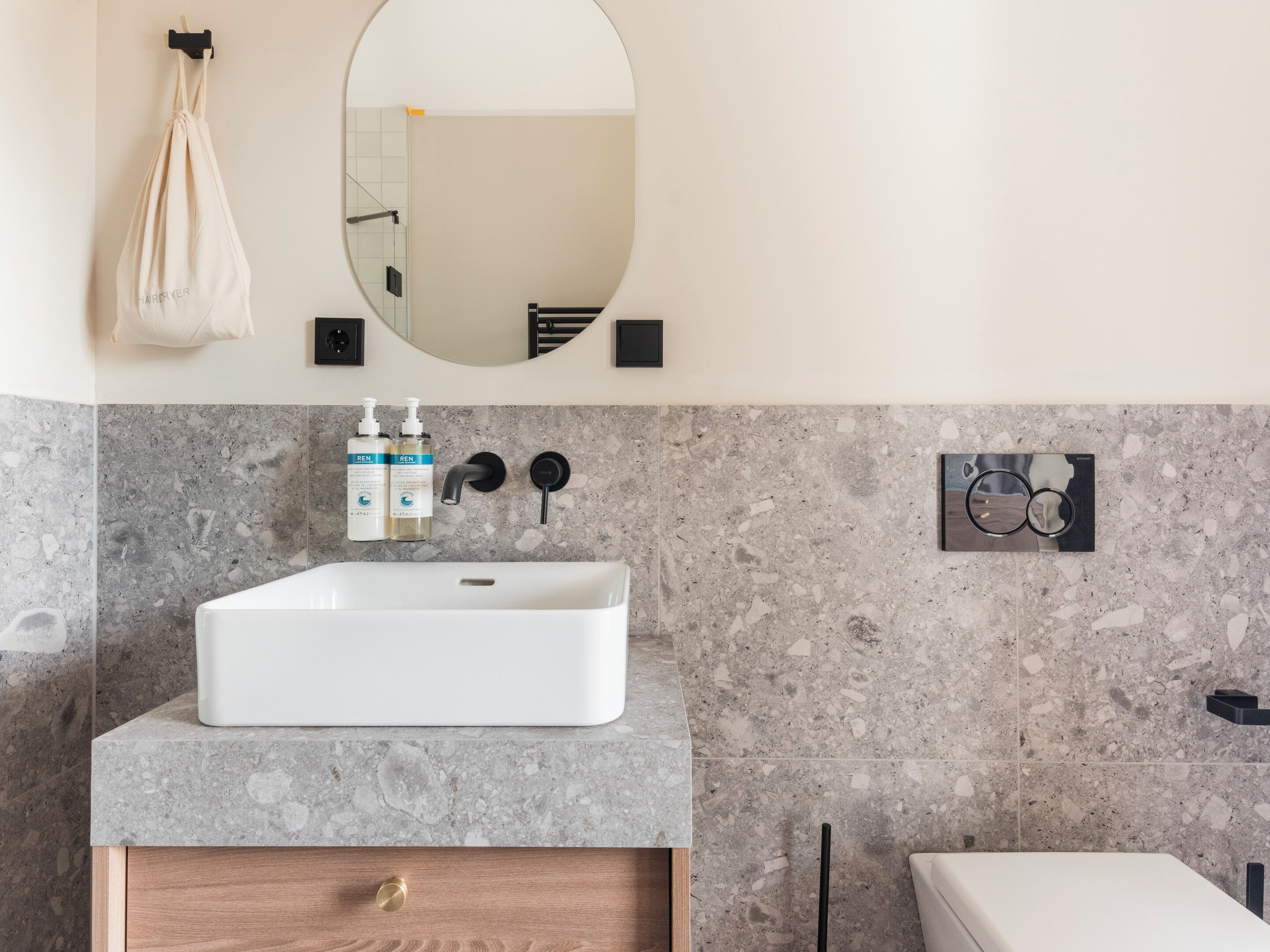 A bathroom with a white sink, marble walls, a wooden cabinet, a mirror, and toiletries.