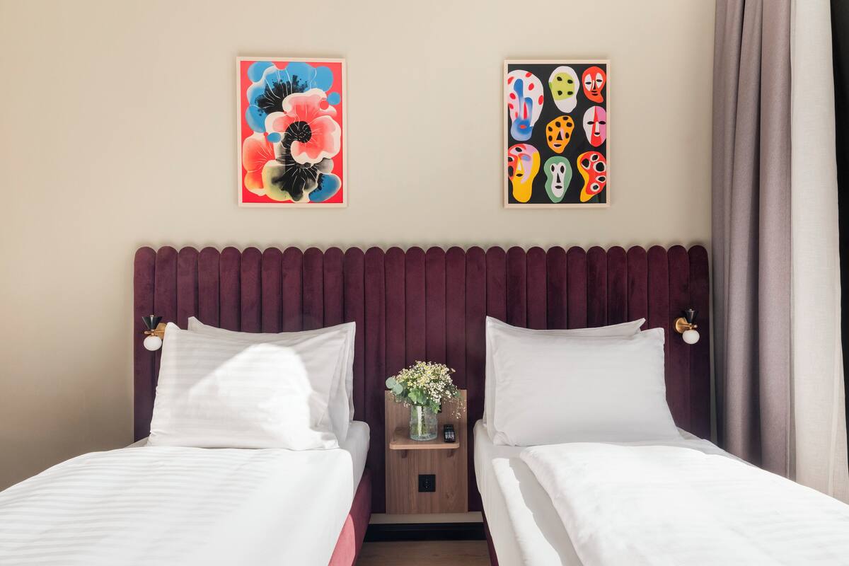 Une chambre d’hôtel avec deux lits, une table de chevet en bois ornée d’un vase de fleurs et deux tableaux accrochés au mur.