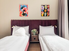 Habitación de hotel con dos camas, una mesita de noche de madera con un jarrón de flores y dos cuadros colgados en la pared.