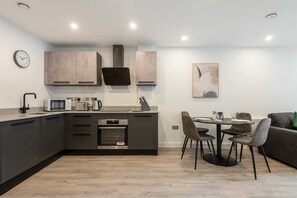 A modern kitchen with dark cabinetry, a microwave, a kettle, and a built-in oven. There is a dining area with a table and chairs. - Stay Zeni, Ouse Court York Stunning 1 Bed Flat (York)