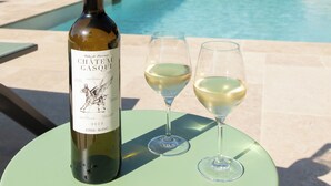 Een fles Chateau Gasqui wijn en twee glazen op een tafel, met een zwembad, ligbedden en parasols op de achtergrond.