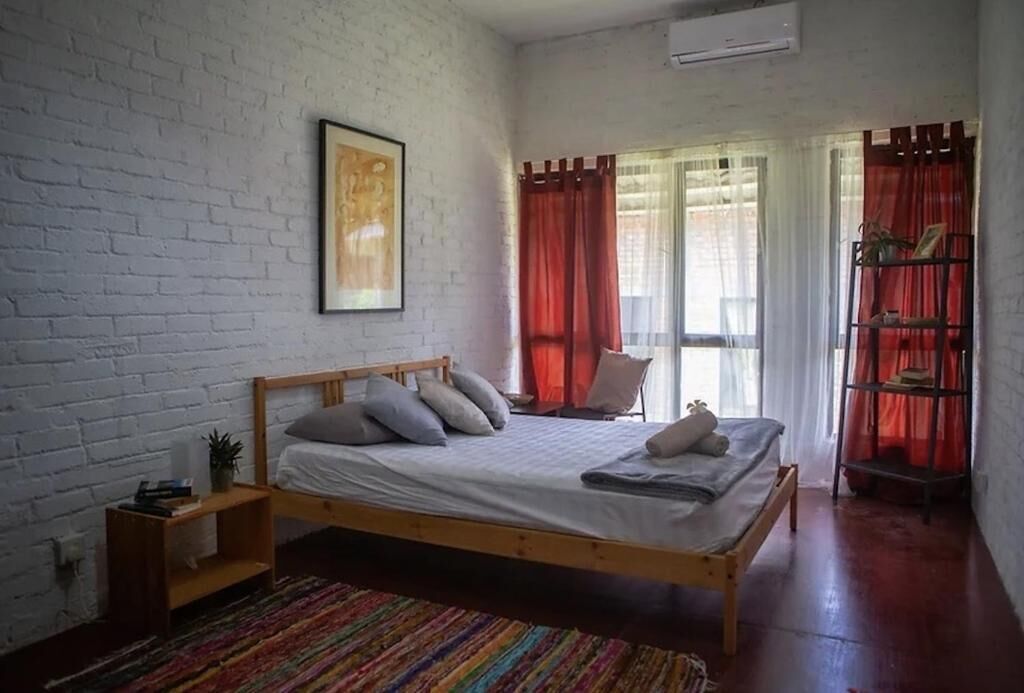 Un dormitorio con una cama de madera, una pared de ladrillo blanco, una alfombra colorida y una estantería con cortinas rojas.