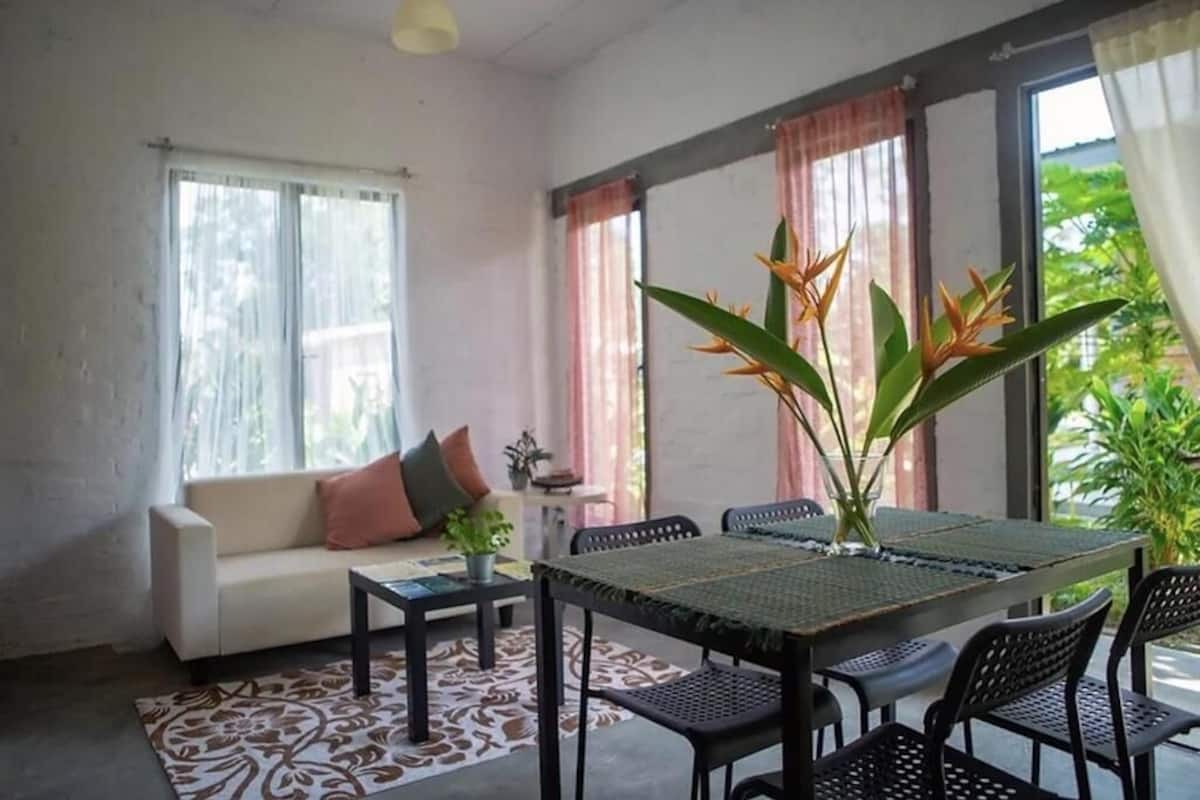 Uma sala de estar com sofá, mesa de centro e mesa de jantar com cadeiras. Há uma janela grande com cortinas e uma planta na mesa.