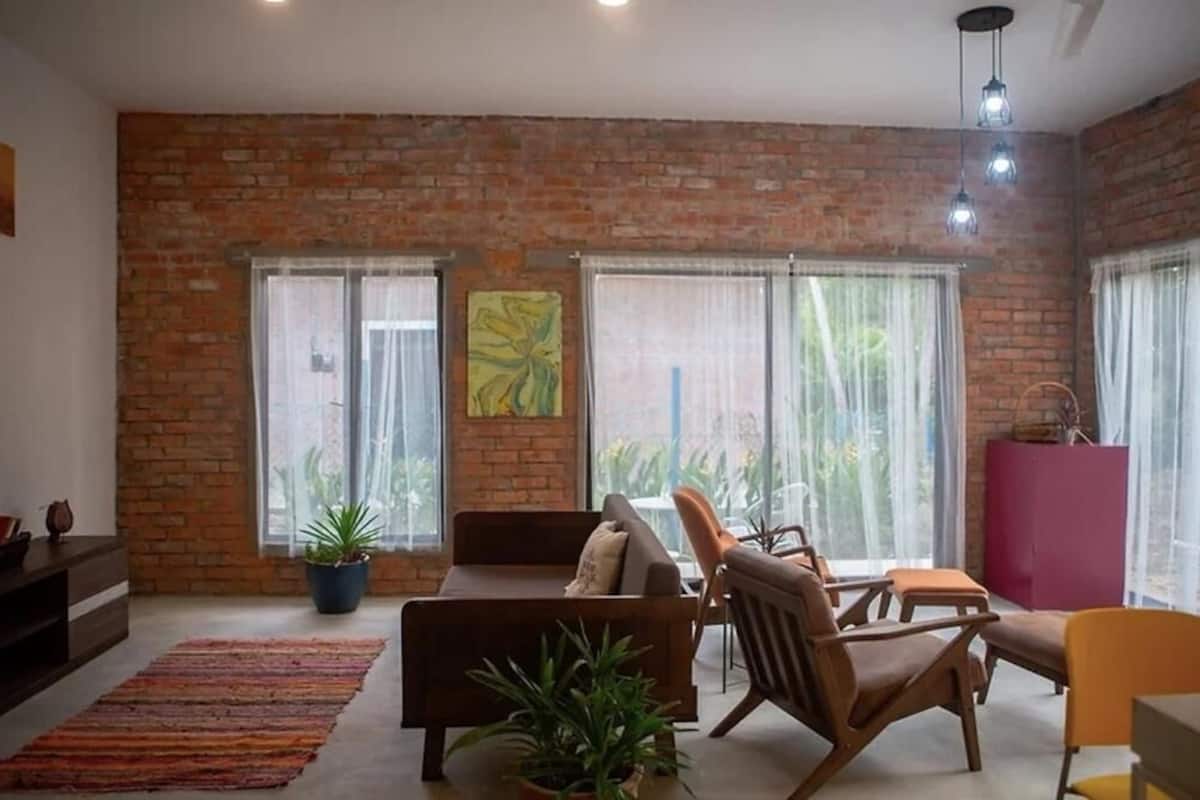 Uma sala de estar com parede de tijolos, janelas amplas, um sofá, cadeiras e uma mesa pequena.