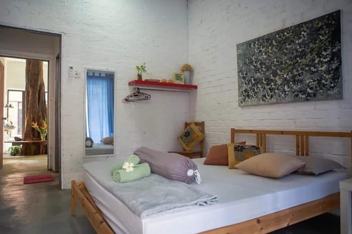 Quarto com cama de madeira, parede de tijolos brancos, uma pintura e uma pequena estante com objetos decorativos.