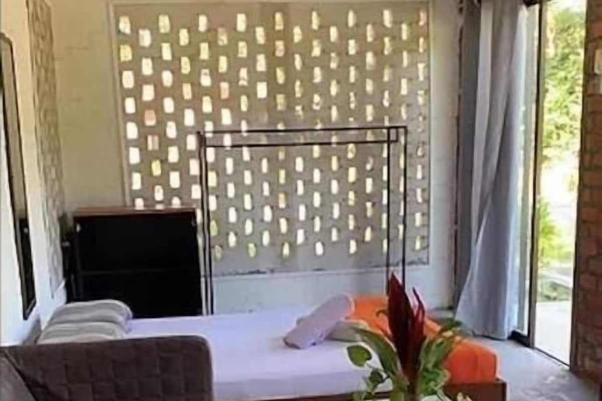 Quarto moderno com cama, uma mesinha e uma cadeira. Possui uma janela com cortinas e vista para o verde lá fora.