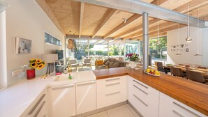 Une cuisine moderne avec des armoires blanches, un comptoir en bois et un grand îlot central. On y trouve un coin repas avec une table et des chaises en bois, ainsi qu’une grande fenêtre qui laisse entrer la lumière naturelle.