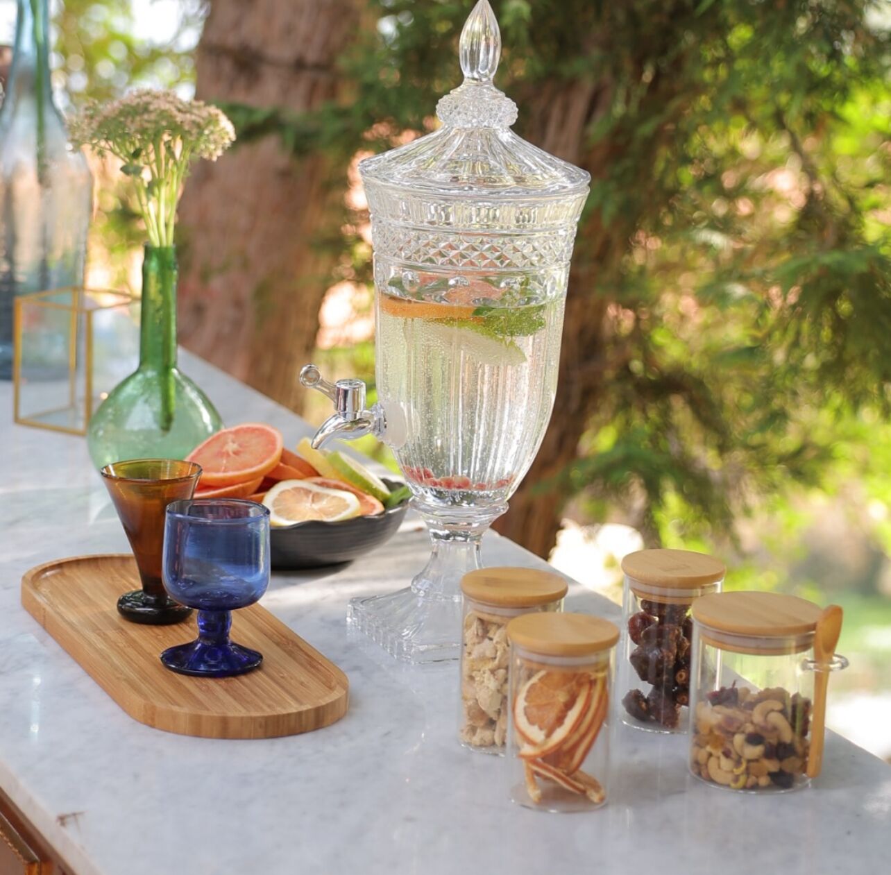 A clear glass punch bowl with a handle, surrounded by various glass jars with wooden lids on a marble countertop.