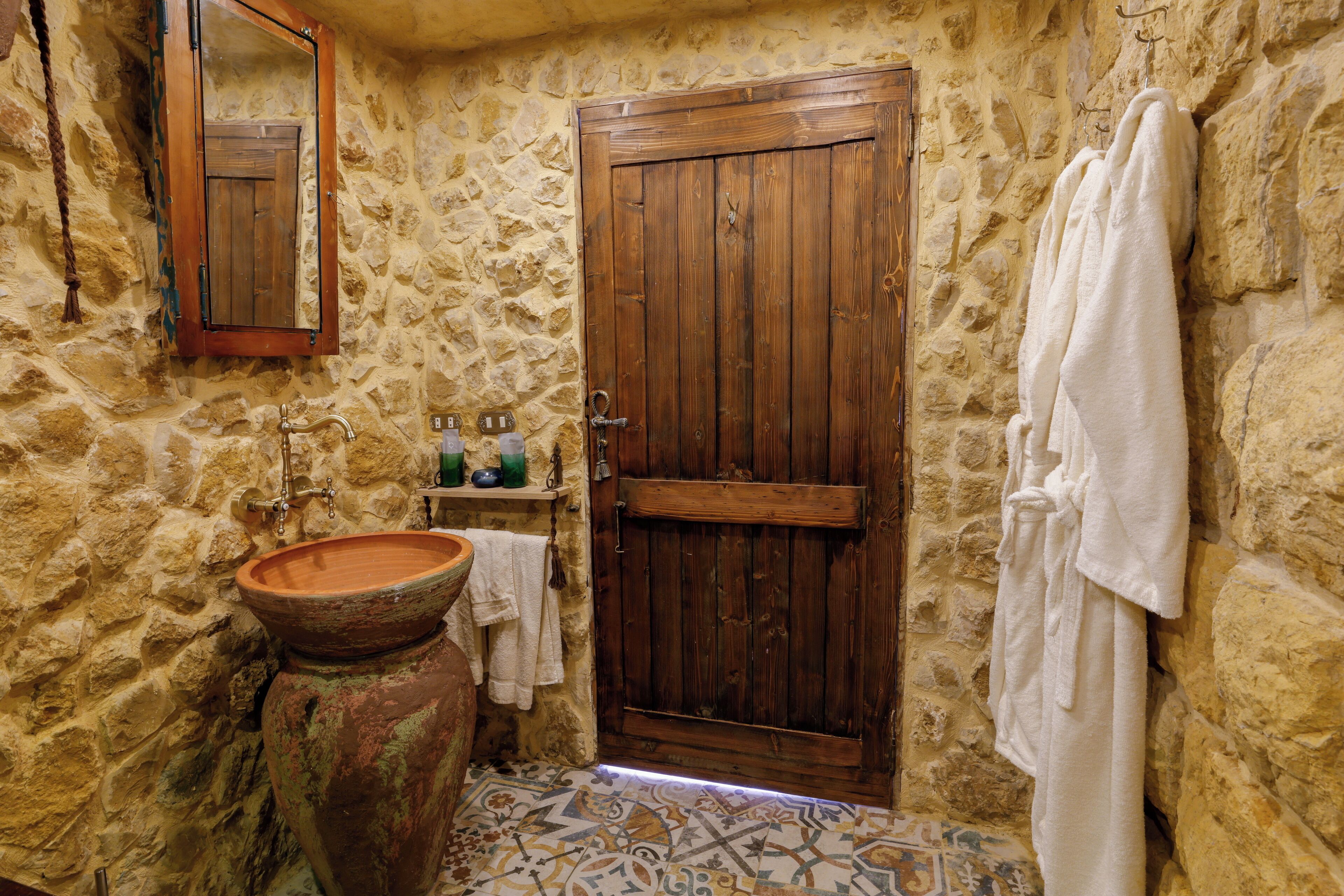 Un baño rústico con pared de piedra, puerta de madera, un lavamanos grande de cerámica y una toalla colgada.