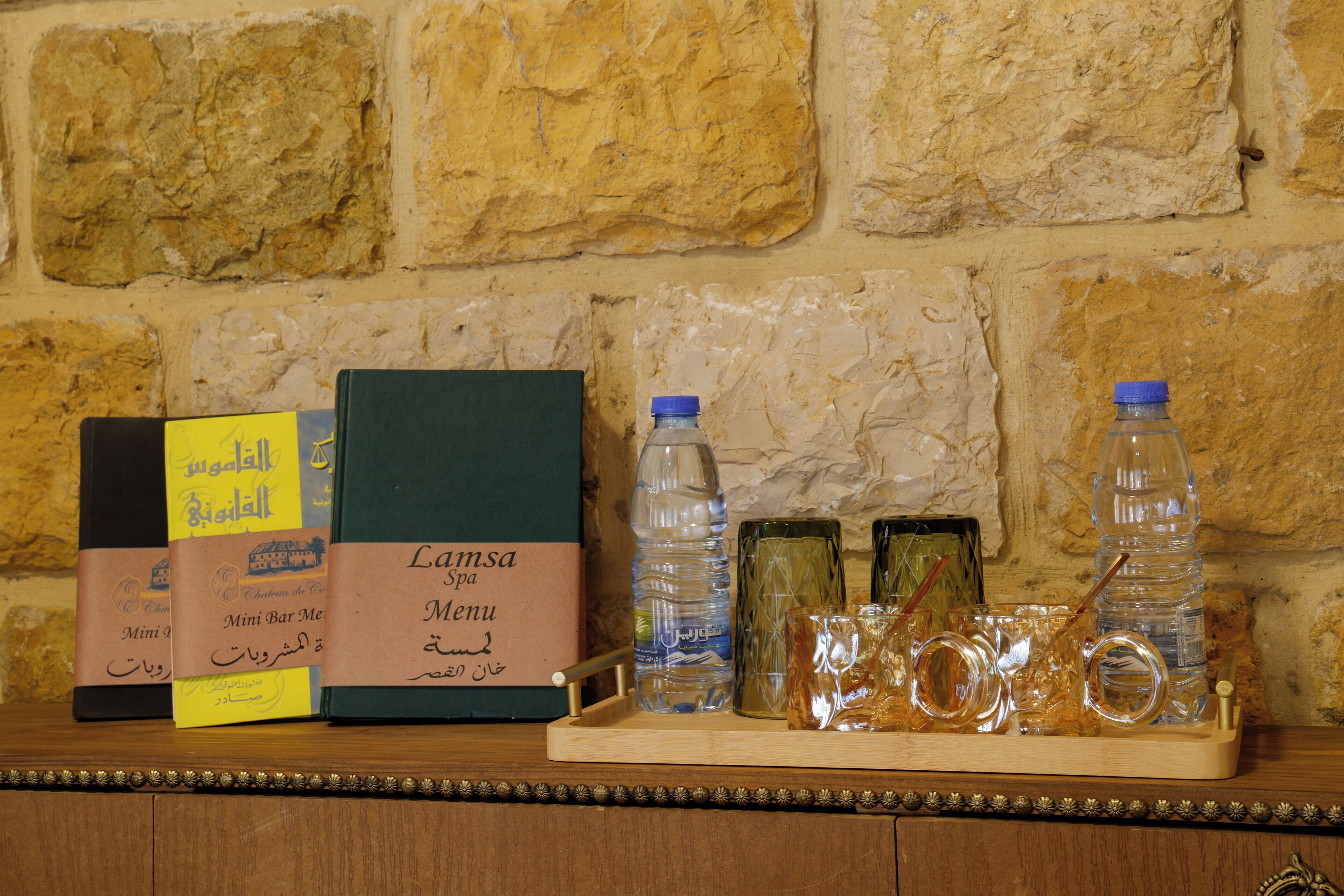 A wooden tray with a menu, water bottles, and decorative glassware on a stone wall background.