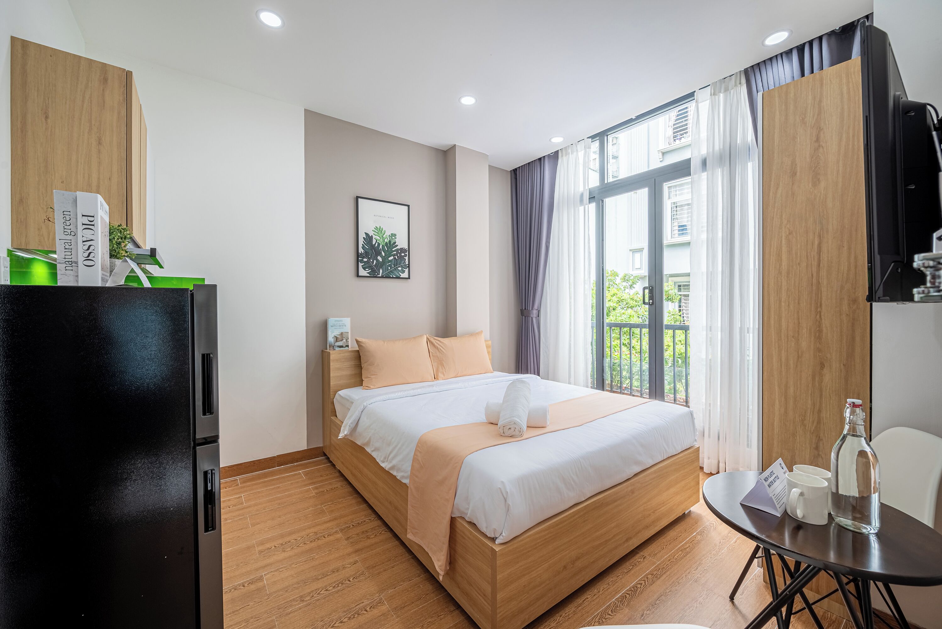 A modern hotel room with a wooden bed, a dining table, a refrigerator, and a view of the outdoors.