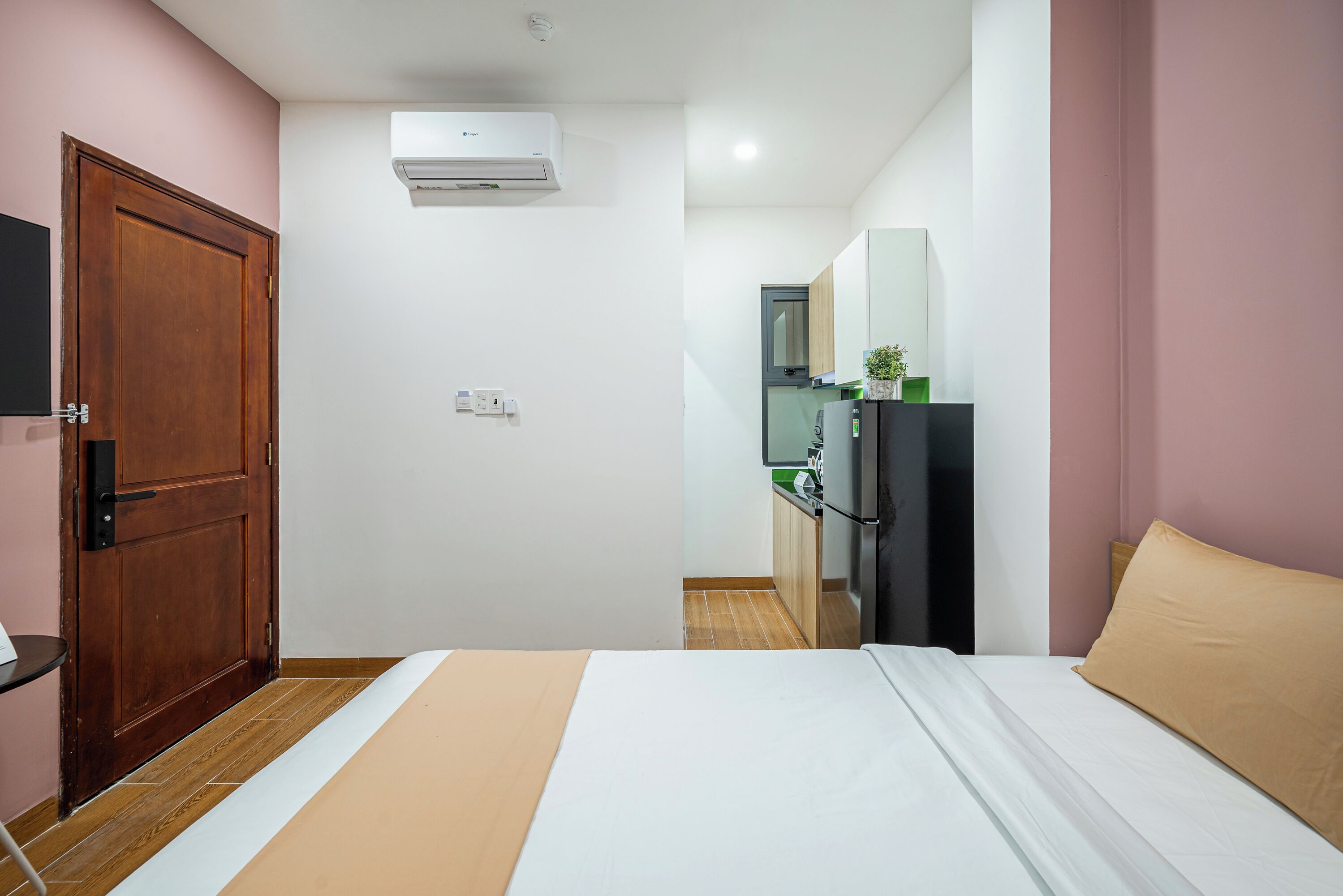 A hotel room with a wooden door, a bed with a beige and white bedspread, a small table, and a kitchenette in the background.